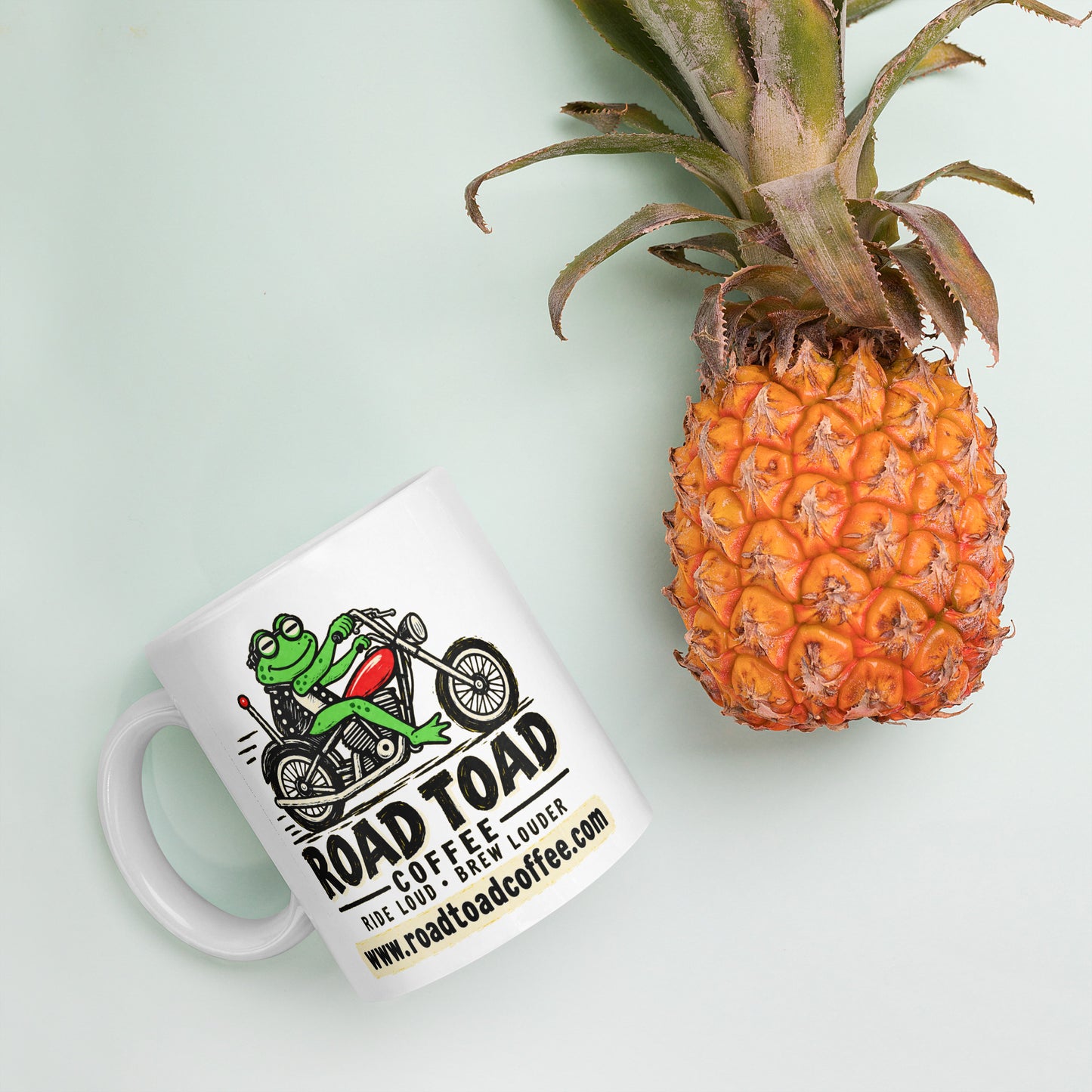 White 11 oz Road Toad Coffee mug with roadtoadcoffee.com next to a pineapple